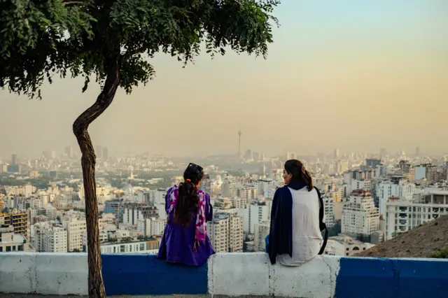 "Tienes que traer la autorización firmada de tu esposo": las duras leyes que enfrentan las mujeres de Irán para conseguir un trabajo 3 Mujeres iraníes sin hiyab obligatorio sentadas con vistas a la ciudad de Teherán, 2024.