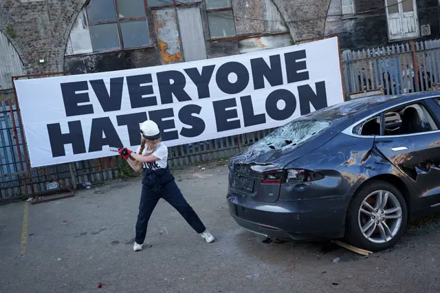 Una mujer golpeando un Tesla con un martillo, con un gran letrero atrás que dice "Todo el Mundo odia a Elon".