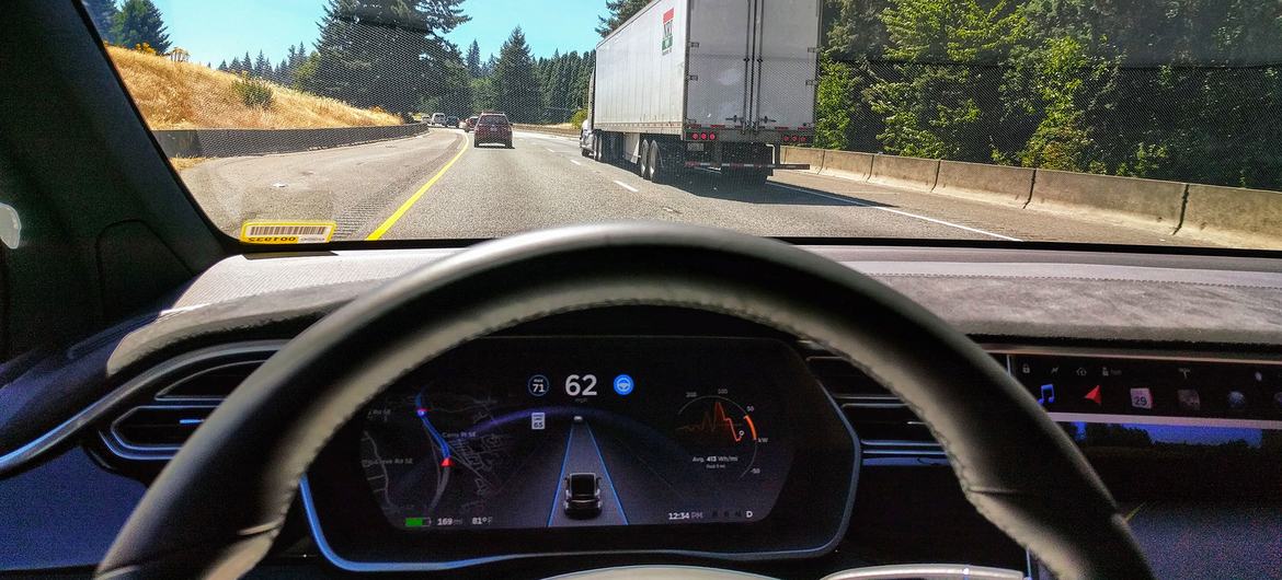 Vista desde el asiento del conductor de un Tesla, que muestra el tablero de instrumentos y una escena de autopista con un camión grande en el carril adyacente.